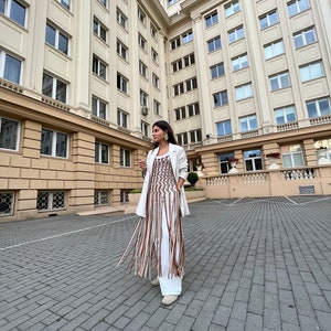 May include: A woman wearing a white blazer and a brown and white fringe top, standing in front of a building with many windows.