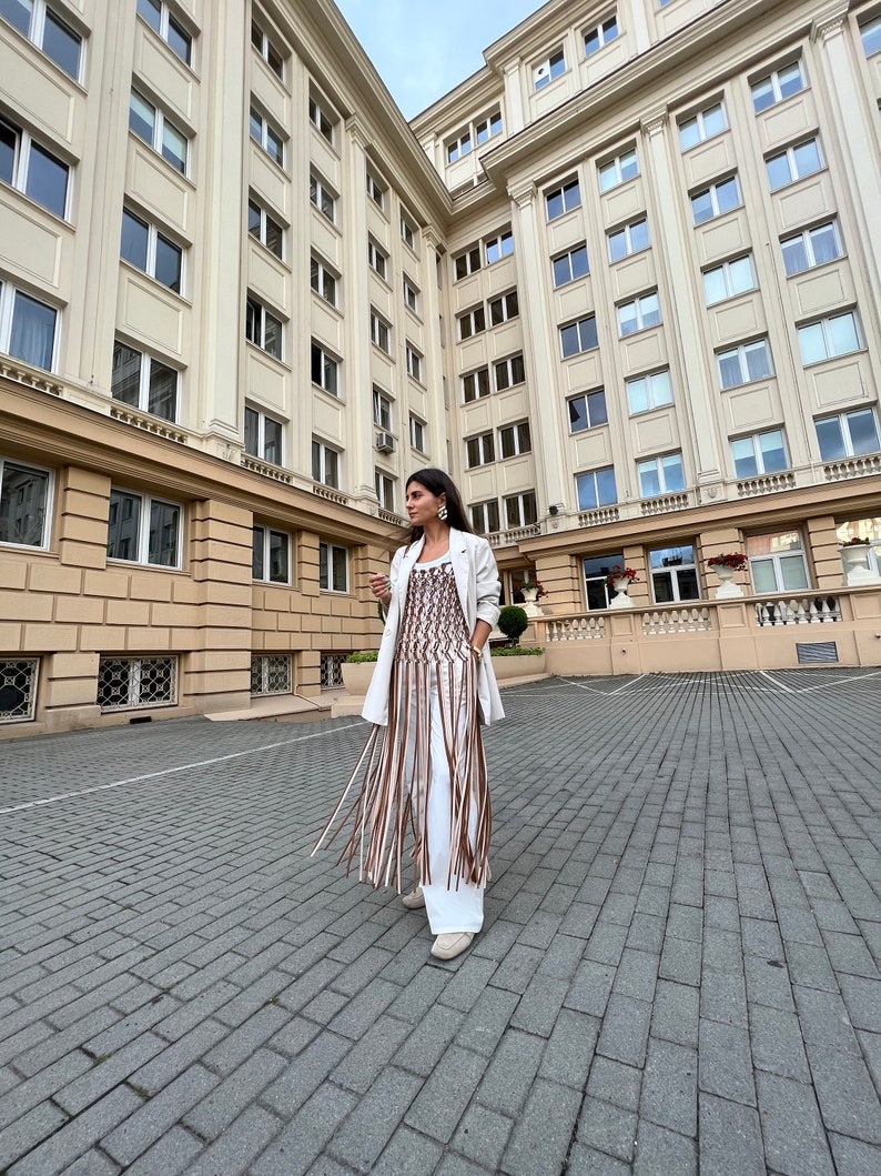 May include: A woman wearing a white blazer and a brown and white fringe top, standing in front of a building with many windows.