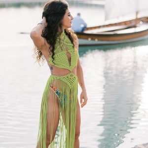 May include: A woman wearing a lime green crochet top and skirt with fringe. She is standing on a stone jetty by a body of water with a sailboat in the background.
