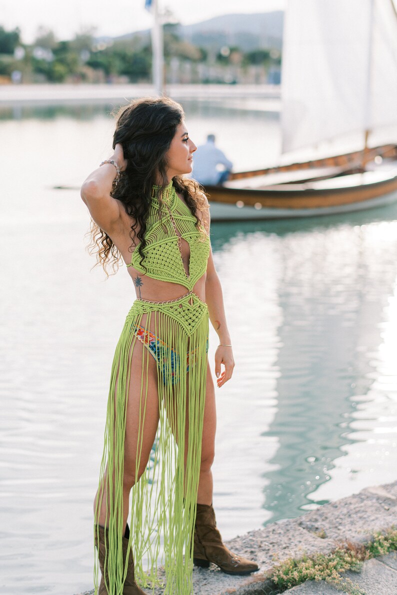 May include: A woman wearing a lime green crochet top and skirt with fringe. She is standing on a stone jetty by a body of water with a sailboat in the background.