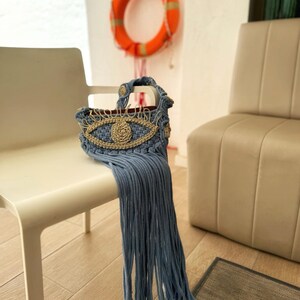 May include: A light blue crocheted bag with a gold evil eye design and long fringe. The bag is sitting on a white chair.