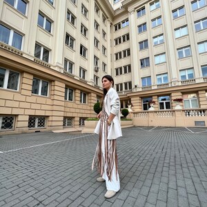 May include: A woman wearing a white blazer and a white dress with brown fringe detail stands in a courtyard. The building behind her has many windows.