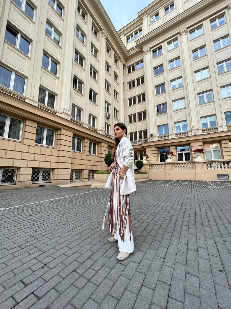 May include: A woman wearing a white blazer and a white dress with brown fringe detail stands in a courtyard. The building behind her has many windows.