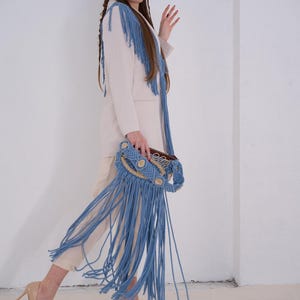 May include: A woman wearing a white blazer with blue fringe trim and light blue fringe pants. She is holding a light blue macrame bag with a brown leather strap.