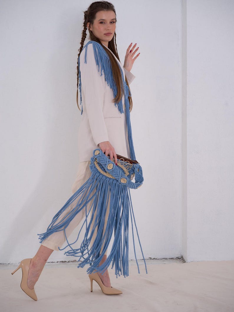 May include: A woman wearing a white blazer with blue fringe trim and light blue fringe pants. She is holding a light blue macrame bag with a brown leather strap.
