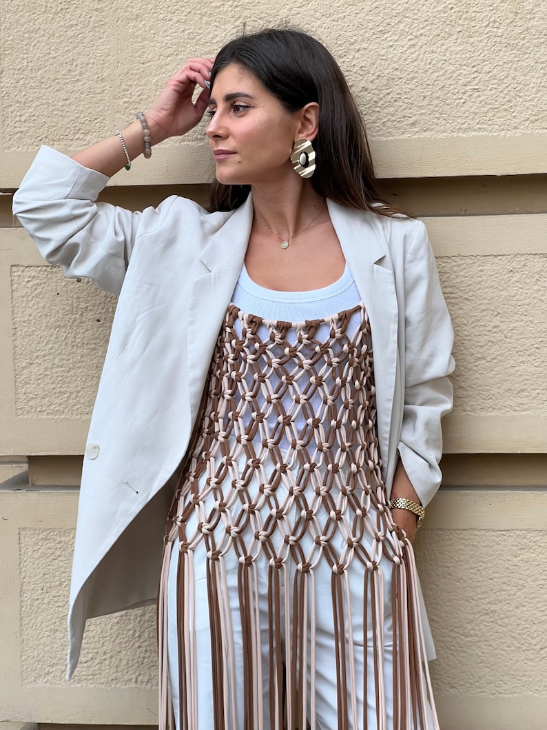 May include: A woman wearing a white blazer over a brown and cream crocheted top with long fringe. The top is worn over a white vest top. She is wearing a gold watch and gold earrings.