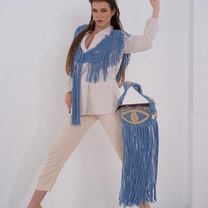 May include: A woman wearing a white blazer and beige pants with a blue crocheted fringe top and matching blue crocheted fringe bag with an eye design.