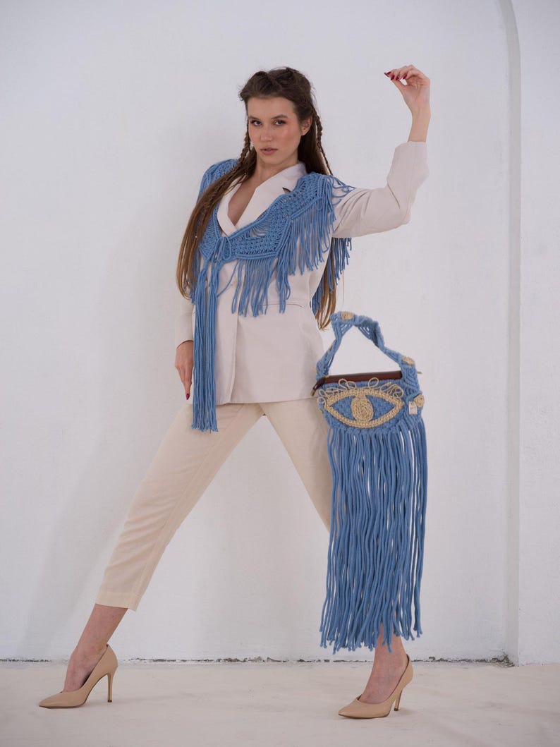 May include: A woman wearing a white blazer and beige pants with a blue crocheted fringe top and matching blue crocheted fringe bag with an eye design.