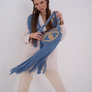 May include: A blue crocheted bag with a large eye design and fringe. The bag is being held by a person wearing a white blazer and beige pants.