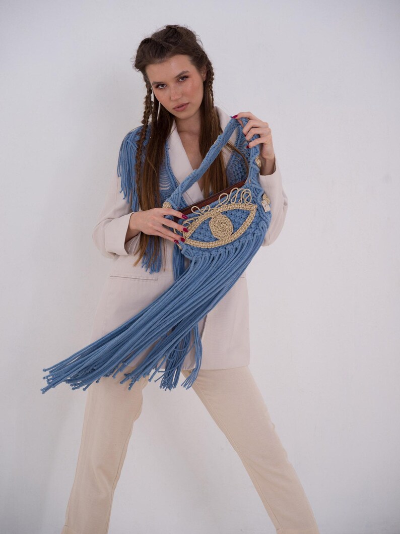 May include: A blue crocheted bag with a large eye design and fringe. The bag is being held by a person wearing a white blazer and beige pants.