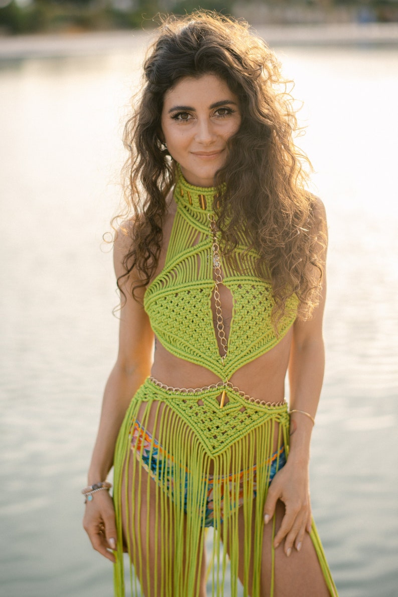 May include: A woman wearing a green crocheted halter top and skirt with fringe. The top features a gold chain detail and the skirt has a gold chain belt. The woman is standing in front of a body of water.