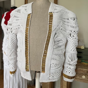 May include: White crocheted jacket with gold accents. The jacket has a geometric pattern and is open in the front.
