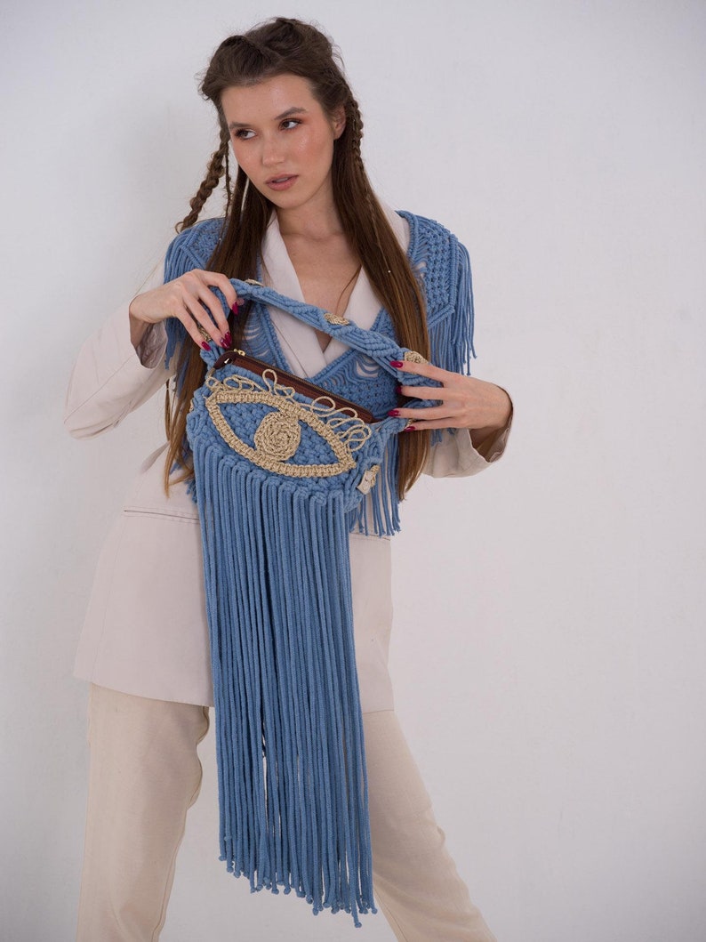 May include: A blue crocheted bag with a large eye design and fringe. The bag is held by a person wearing a beige blazer and pants.
