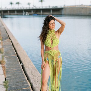 May include: A woman wearing a lime green crochet halter top and skirt with fringe. She is standing on a concrete ledge by a body of water.