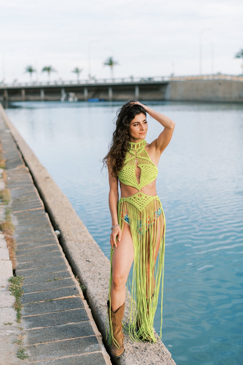 May include: A woman wearing a lime green crochet halter top and skirt with fringe. She is standing on a concrete ledge by a body of water.