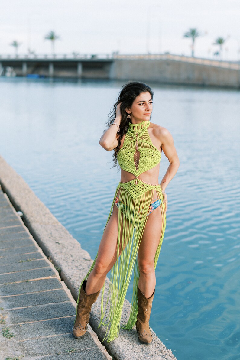 May include: A woman wearing a lime green crochet halter top and skirt with fringe. She is standing on a concrete pier by the water. She is wearing brown cowboy boots.
