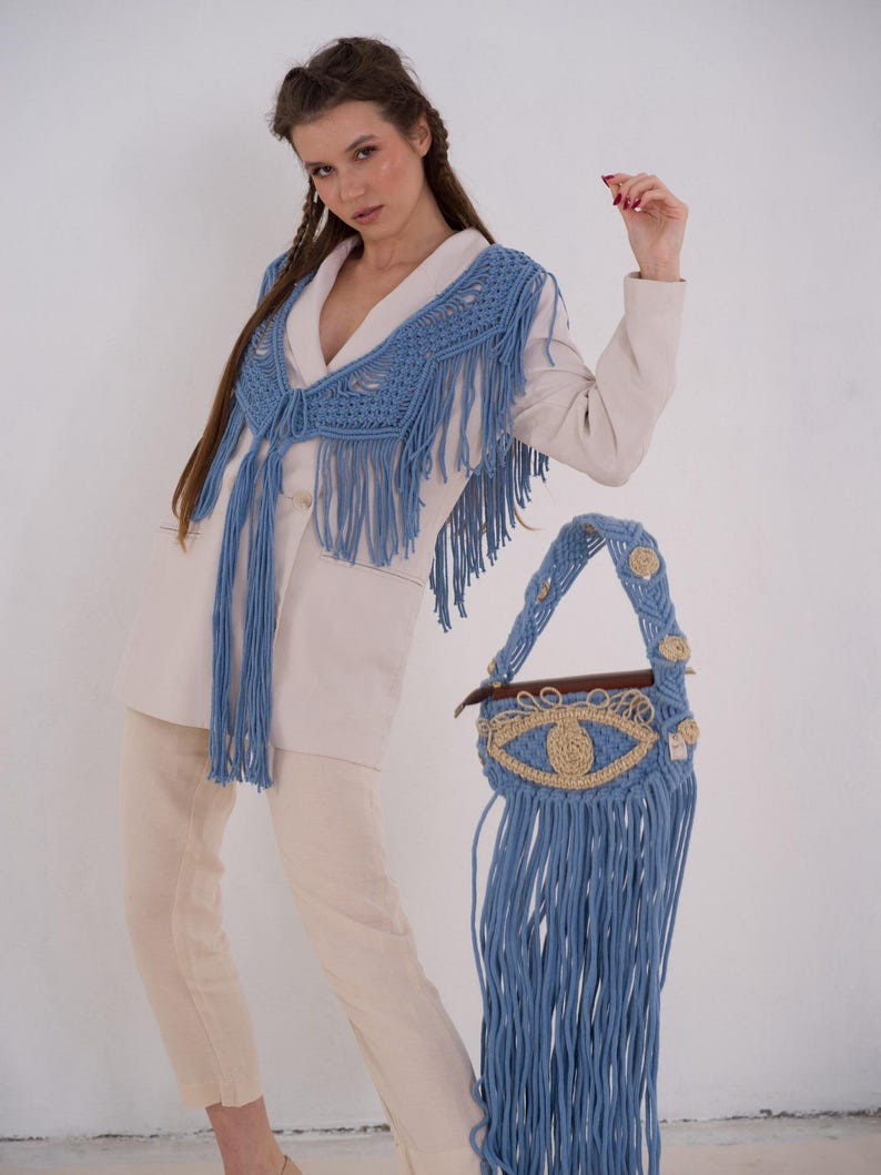 May include: A woman wearing a white blazer and cream pants with a blue crocheted fringe shawl and matching blue crocheted fringe bag with an eye design.