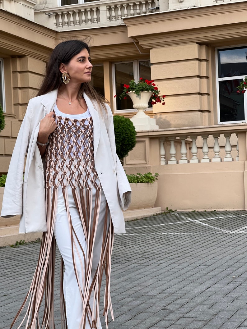 May include: A woman wearing a white blazer over a brown and white crocheted top with long fringe. The top is a sleeveless, fitted style. The woman is standing in front of a building with a stone facade.