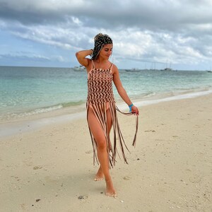 May include: A woman wearing a brown and white crochet cover-up with fringe on a sandy beach.