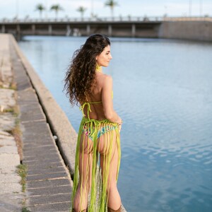 May include: A woman wearing a green crochet bikini top and skirt with fringe. She is standing on a concrete pier by a body of water. She is wearing brown cowboy boots.