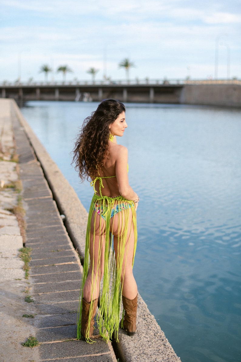 May include: A woman wearing a green crochet bikini top and skirt with fringe. She is standing on a concrete pier by a body of water. She is wearing brown cowboy boots.