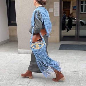May include: A woman wearing a gray blazer with a blue fringed shawl and a blue and gold eye-shaped crocheted bag.