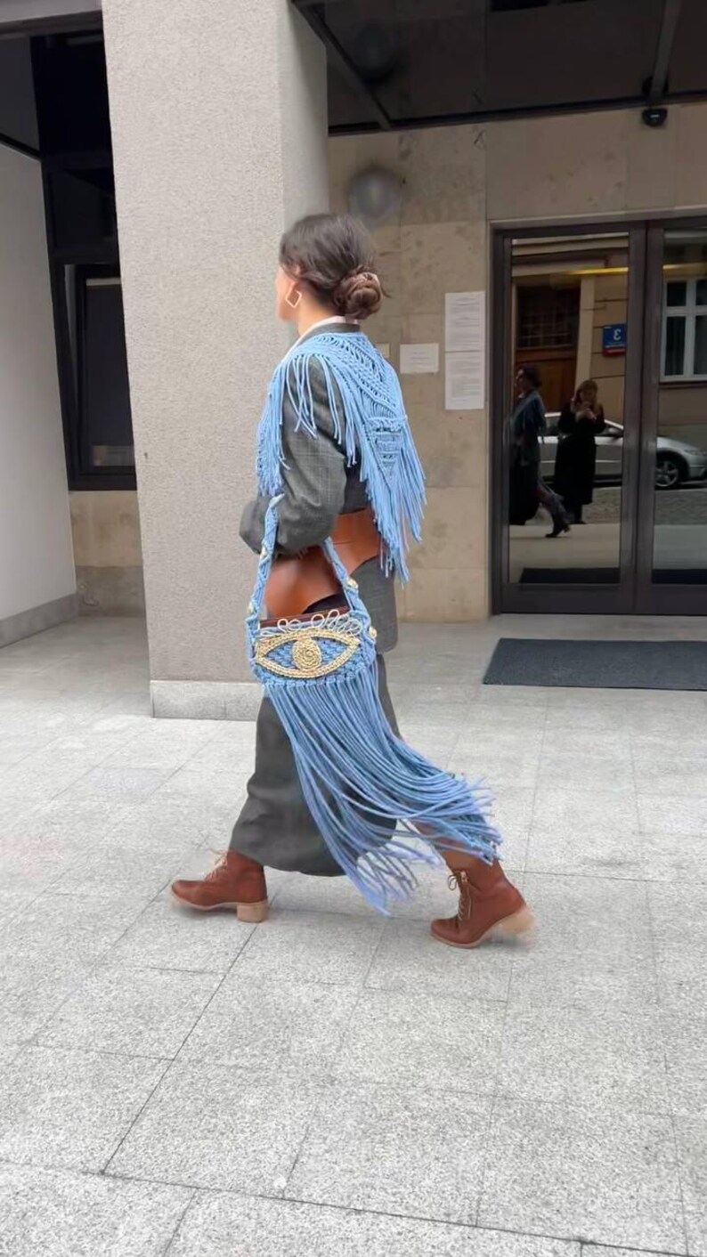 May include: A woman wearing a gray blazer with a blue fringed shawl and a blue and gold eye-shaped crocheted bag.