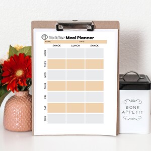 May include: A printable toddler meal planner on a clipboard with a white background. The planner has columns for each day of the week and rows for snacks and lunch. The planner has the text "Toddler Meal Planner" at the top.