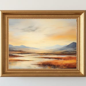 May include: A framed landscape painting with a gold-colored ornate frame. The artwork depicts a serene scene of mountains, a body of water, and a colorful sky with shades of orange, yellow, and blue. The painting is mounted on a white wall.