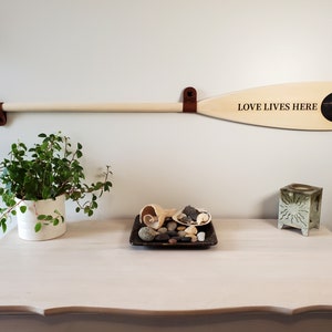 May include: A wooden oar with the text "LOVE LIVES HERE" and a black and white image is mounted on a white wall with brown leather straps. A white table below holds a plant, shells, and a decorative candle holder.