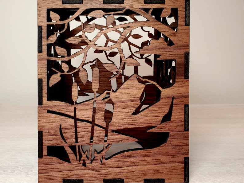 May include: A wooden box with a laser-cut design of cattails and reeds in a pond. The box is made of dark brown wood with black accents.