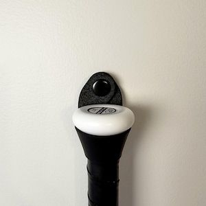 May include: A black baseball bat holder with a felt backing and a black button. The holder is mounted on a white wall.