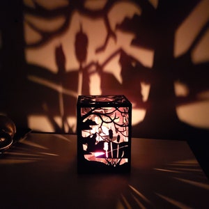 May include: A black metal candle holder with a cut-out design of a forest scene. The candle holder is lit and casting a shadow on the wall behind it.
