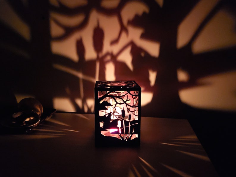 May include: A black metal candle holder with a cut-out design of a forest scene. The candle holder is lit and casting a shadow on the wall behind it.