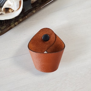 May include: A brown leather cup holder with a black button closure. The cup holder is made of textured leather and has a folded over design.