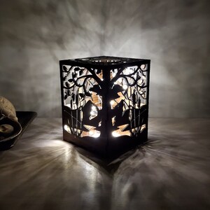 May include: A black wooden lamp with a cutout design of a tree and birds. The lamp is lit and casting a warm glow.