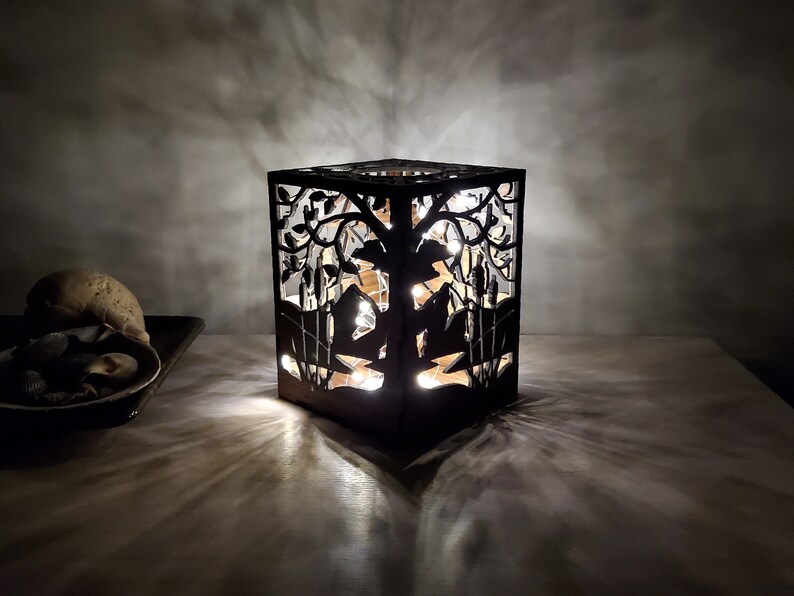 May include: A black wooden lamp with a cutout design of a tree and birds. The lamp is lit and casting a warm glow.