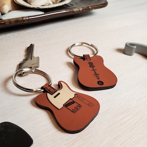 May include: Two guitar-shaped keychains. One is brown with a white pickguard and the other is brown with a fretboard design. Both have silver key rings.