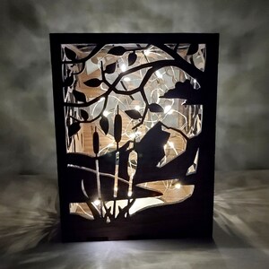 May include: A black wooden lantern with a cut-out design of a tree branch with leaves and cattails. The lantern is illuminated with white fairy lights.