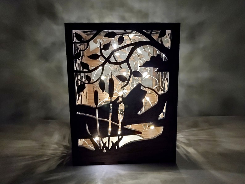 May include: A black wooden lantern with a cut-out design of a tree branch with leaves and cattails. The lantern is illuminated with white fairy lights.
