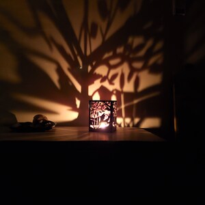 May include: A metal candle holder with a cut-out design of a tree and a house. The candle holder is lit and casting a shadow of the design on the wall behind it.