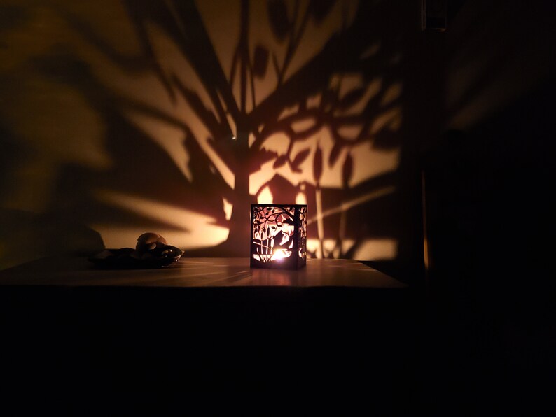 May include: A metal candle holder with a cut-out design of a tree and a house. The candle holder is lit and casting a shadow of the design on the wall behind it.