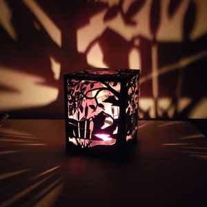May include: A black metal candle holder with a cut-out design of a tree and birds. The candle holder is lit and casting shadows on the wall behind it.