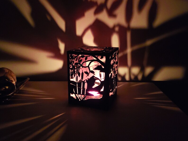 May include: A black metal candle holder with a cut-out design of a tree and birds. The candle holder is lit and casting shadows on the wall behind it.