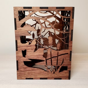 May include: A wooden box with a cutout design of a forest scene. The box is made of dark wood and has a black border. The cutout design features trees, branches, and cattails.