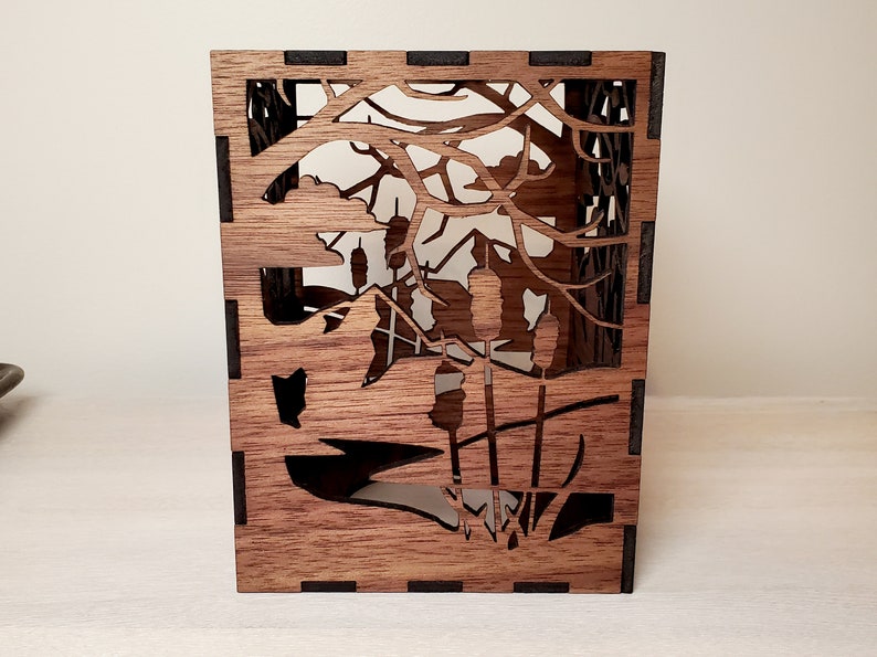 May include: A wooden box with a cutout design of a forest scene. The box is made of dark wood and has a black border. The cutout design features trees, branches, and cattails.