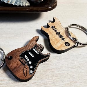 Puede incluir: Dos llaveros de guitarra de madera, uno con un diseño de guitarra en blanco y negro y el otro con un diseño de código de barras en blanco y negro. Ambos llaveros tienen anillas de llavero plateadas.