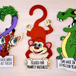Puede incluir: Tres perchas de madera con forma de animales con letreros humorísticos. Una jirafa morada con hojas verdes en la cabeza dice "Vuelve cuando sea un poco más alto". Un mono rojo con un letrero que dice "Cerrado por asuntos de mono". Un dinosaurio verde con un letrero que dice "Vuelve en 20 millones de años".