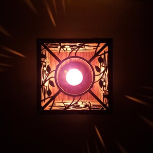 May include: A square, wooden light fixture with a decorative, laser-cut design of leaves and vines. The light fixture has a warm, white light bulb inside.