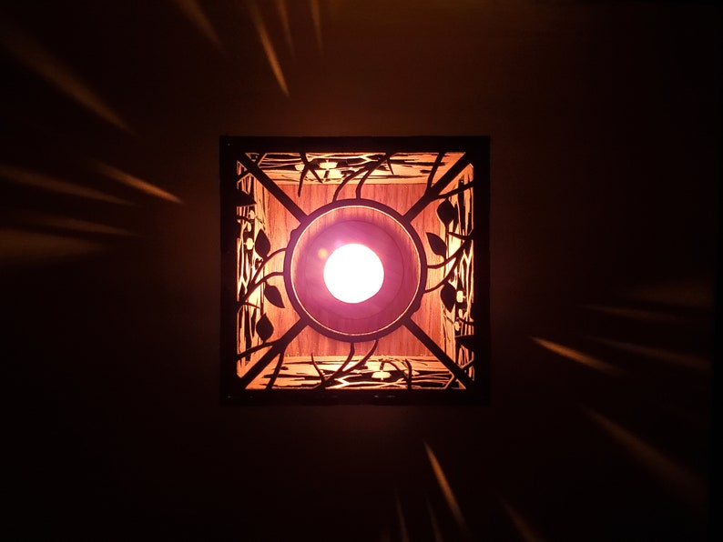 May include: A square, wooden light fixture with a decorative, laser-cut design of leaves and vines. The light fixture has a warm, white light bulb inside.